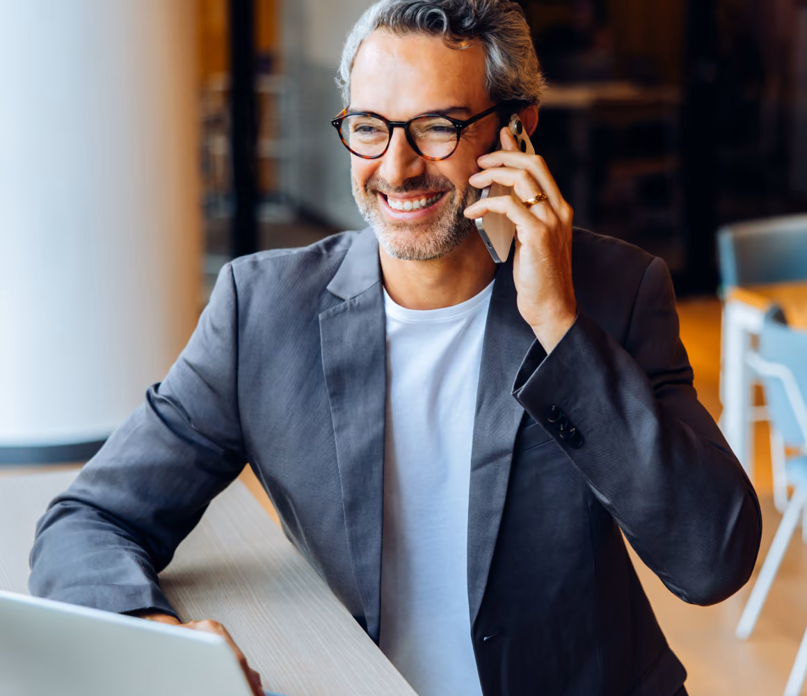 A smiling businessman in a blazer talking on his phone beside an open laptop