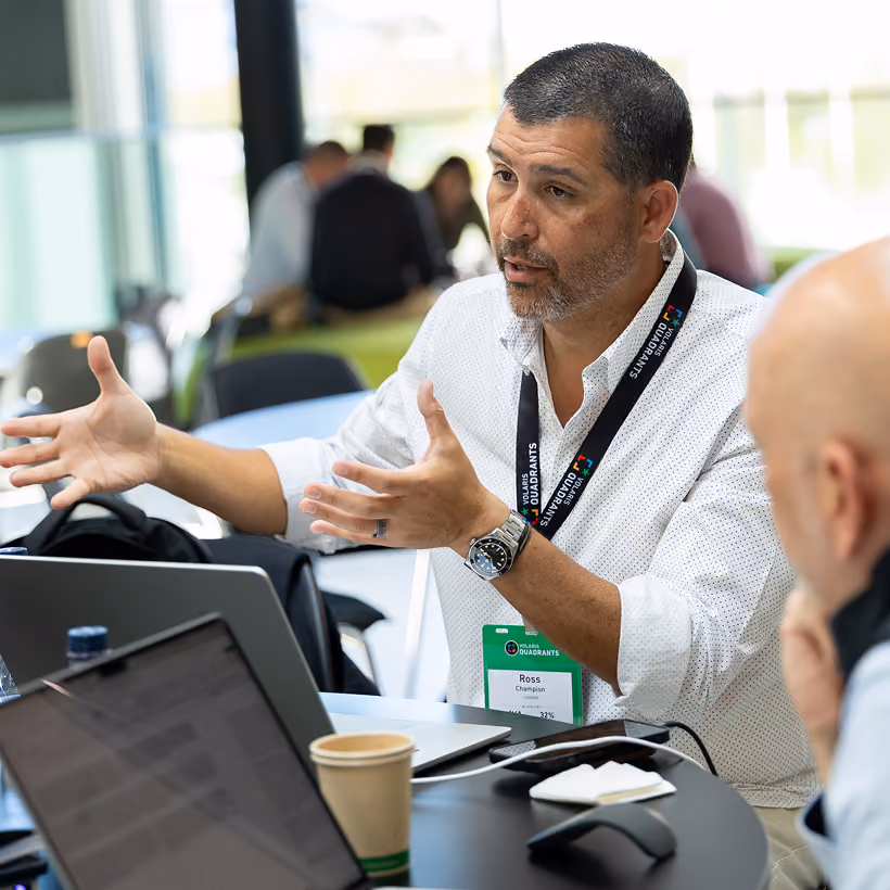 A man gesturing while talking with a colleague at a table with laptops