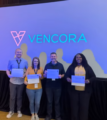 Four colleagues holding certificates on stage in front of a screen showing the Vencora logo