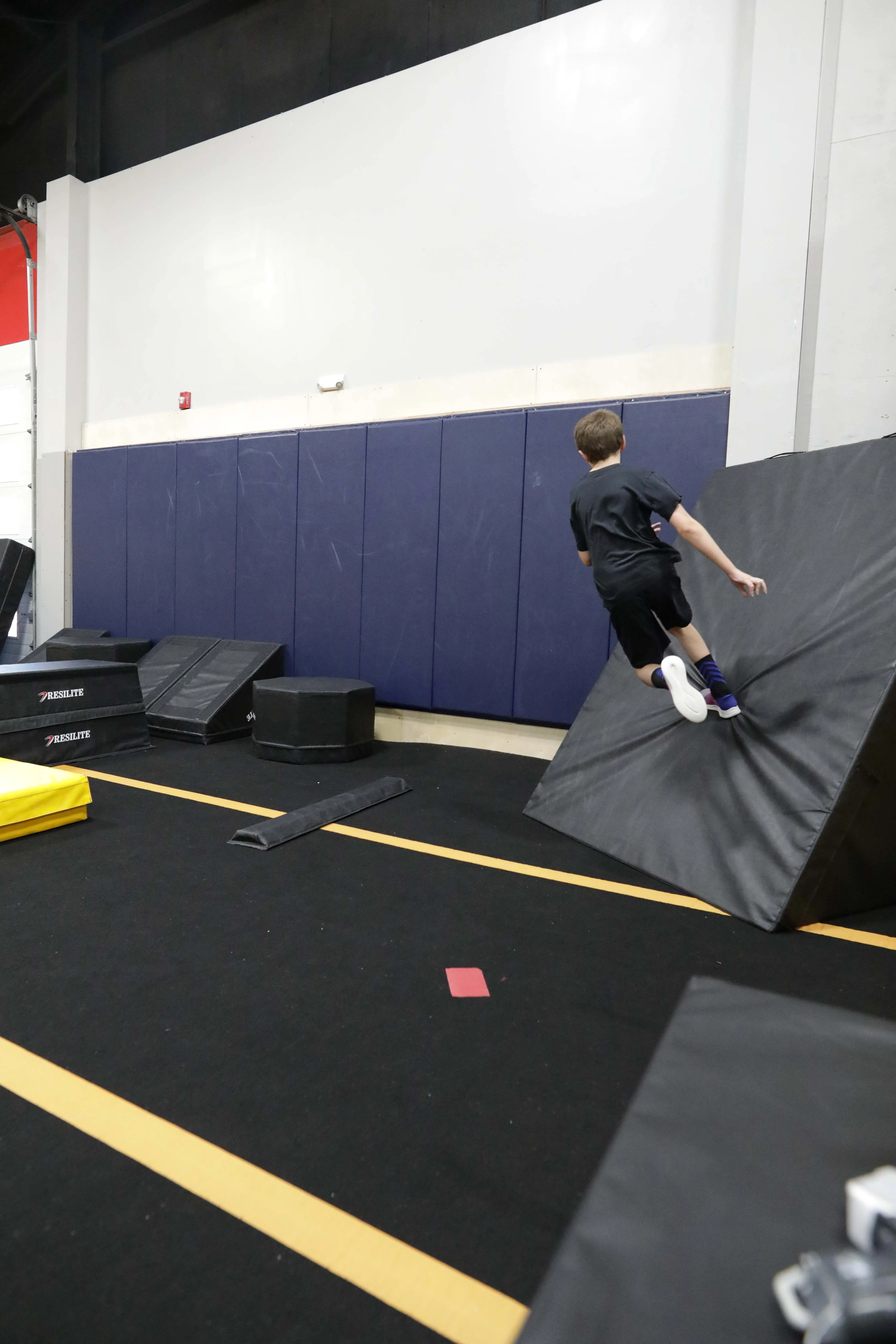 A child practicing a controlled jump over a wooden obstacle with a coach nearby