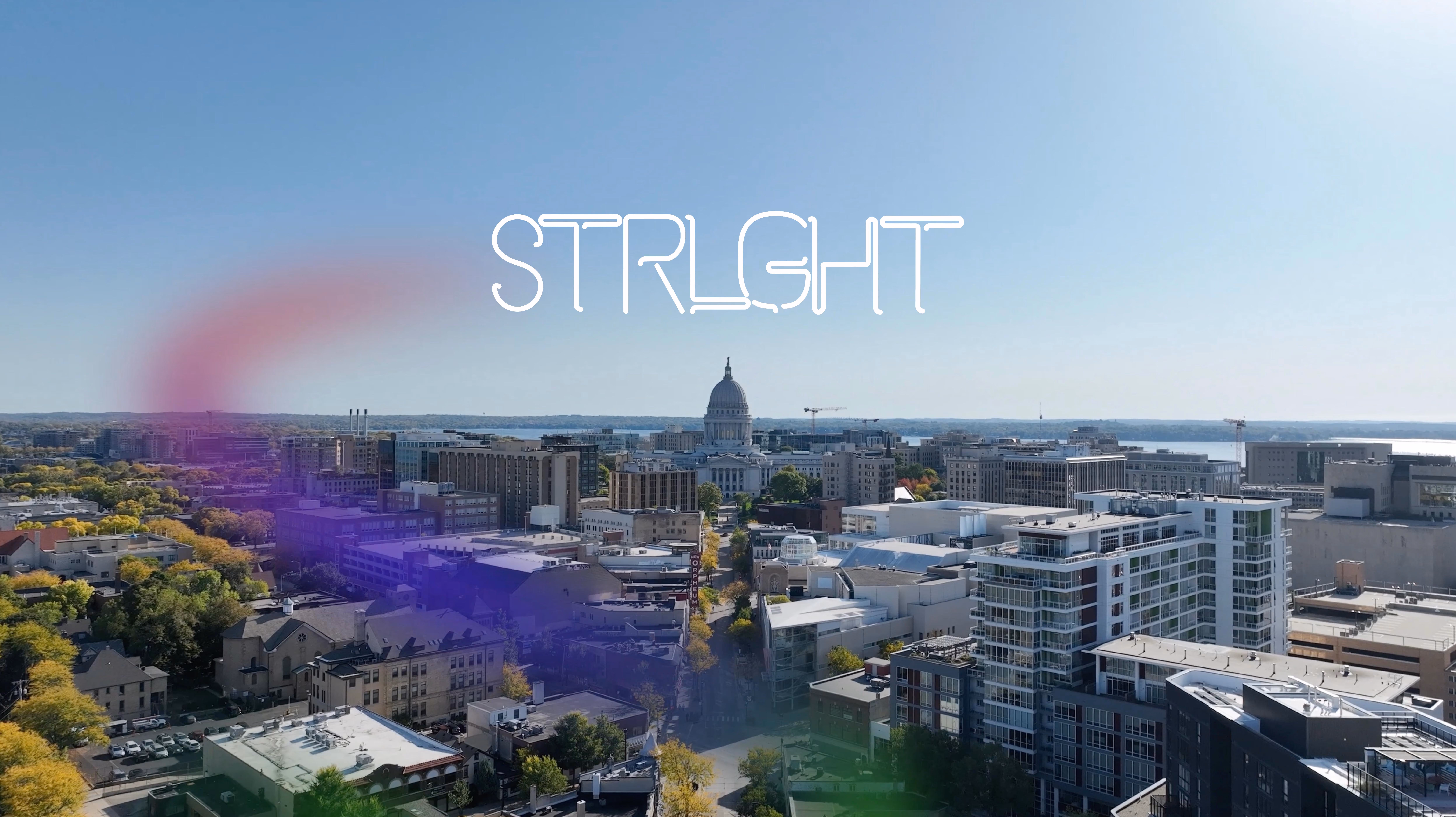 A cityscape with a clear blue sky showing a prominent domed capitol building in the center and modern buildings in the foreground, with the word 'STRLGHT' in stylized white font over the sky and a faint colorful smoke effect towards the left.