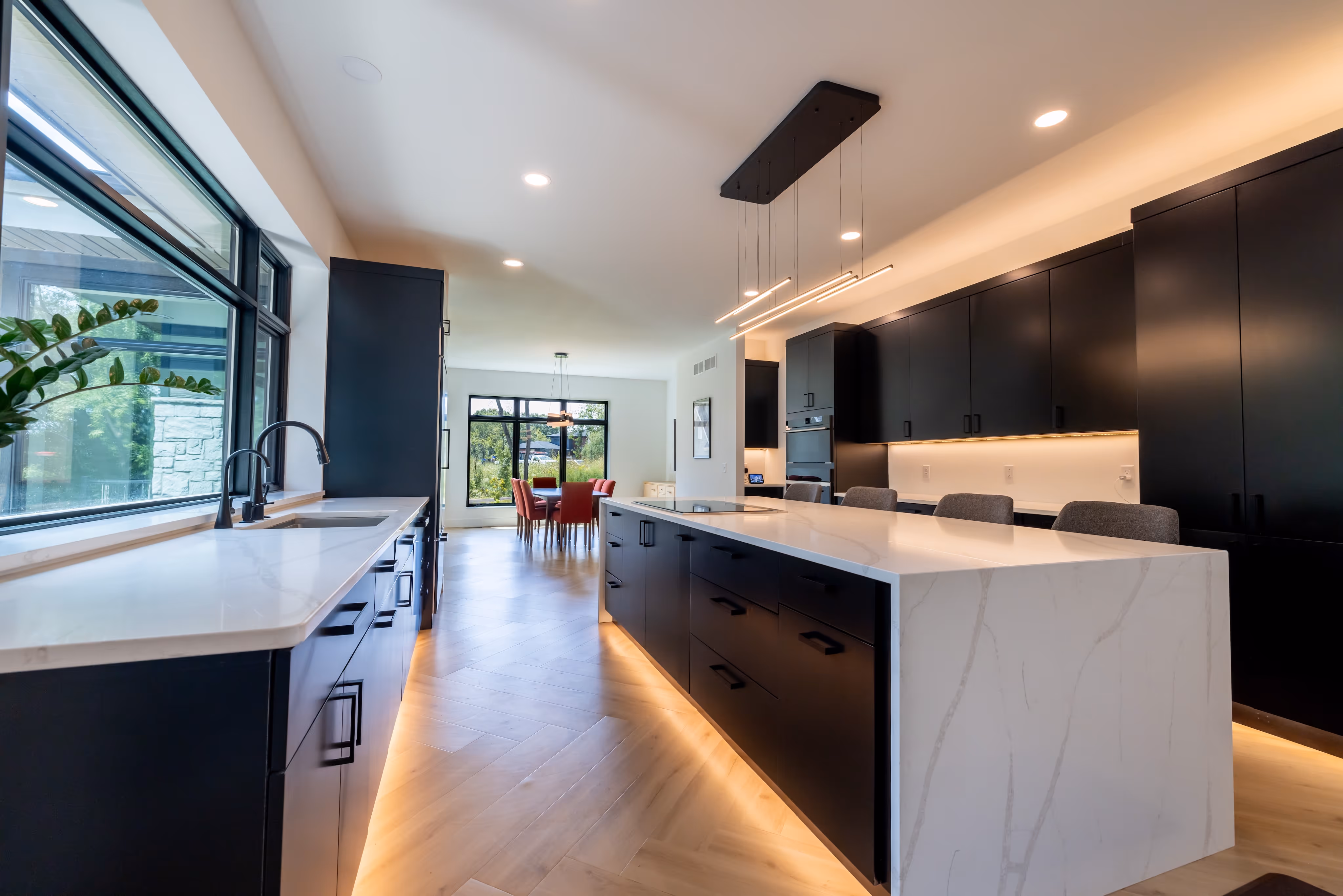 Modern kitchen with large marble island and black cabinetry, illuminated by under-cabinet and pendant lighting, featuring a window with view of greenery and a dining area with red chairs in the background.