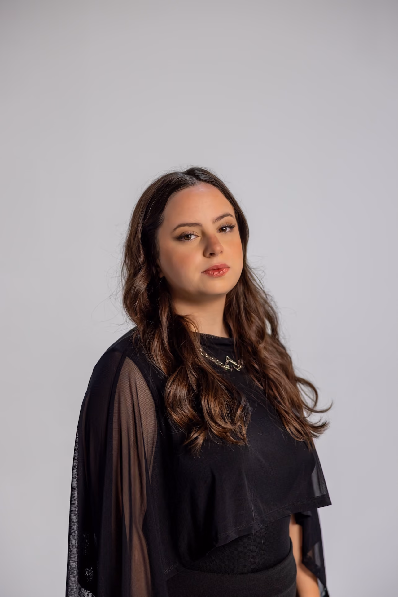 Woman with long wavy brown hair wearing a black top with sheer sleeves, posing against a plain light gray background.