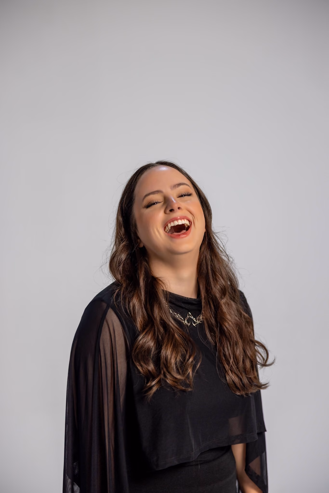 A woman with long wavy brown hair wearing a black outfit and a silver necklace, laughing joyfully against a plain light gray background.