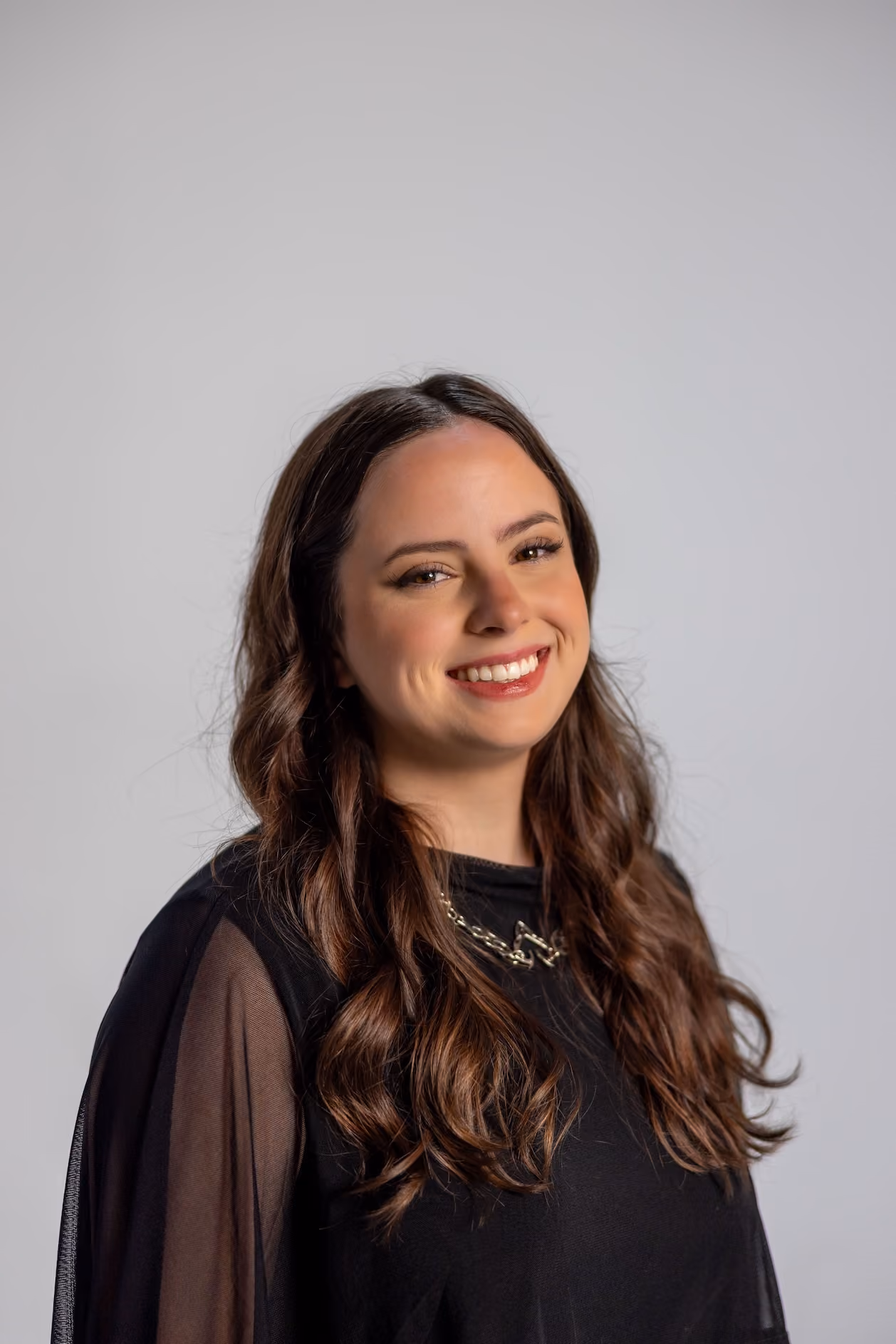 Young woman with long wavy brown hair wearing a black top with sheer sleeves and a gold chain necklace, smiling against a plain gray background.