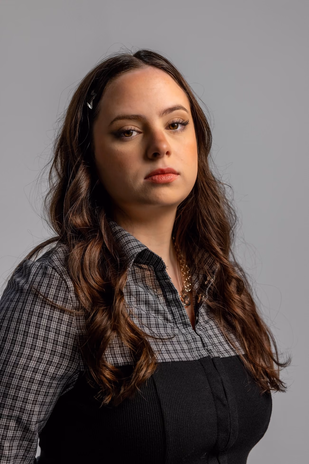 Portrait of a woman with long, wavy brown hair wearing a gray plaid shirt and black top, looking at the camera with a neutral expression against a plain gray background.