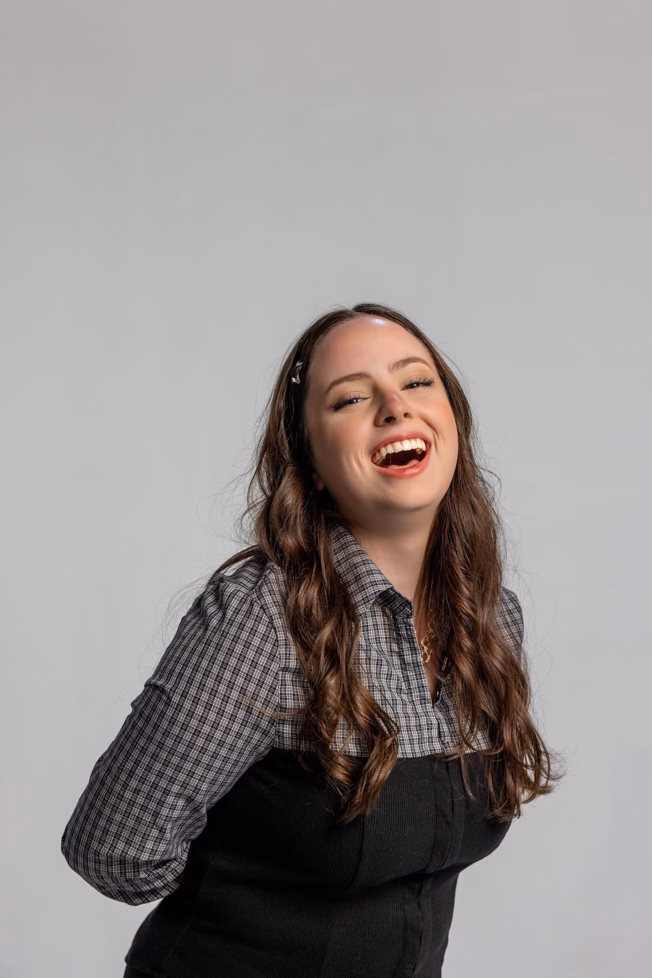 Smiling woman with long wavy brown hair wearing a checkered shirt under a black top, laughing against a gray background.