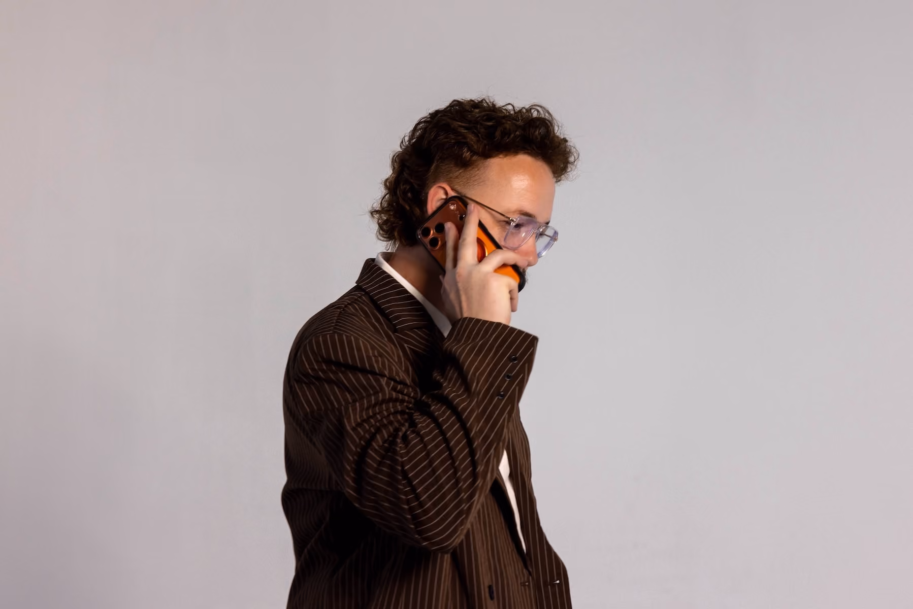 Young man with curly hair and glasses wearing a brown pinstripe blazer talking on an orange smartphone against a plain light background.