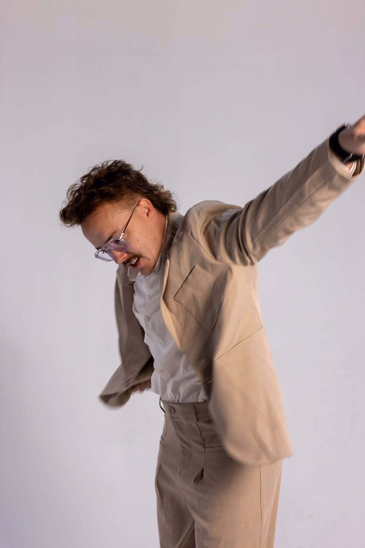 Man wearing beige suit and white shirt with glasses, raising arms and looking down against a plain background.
