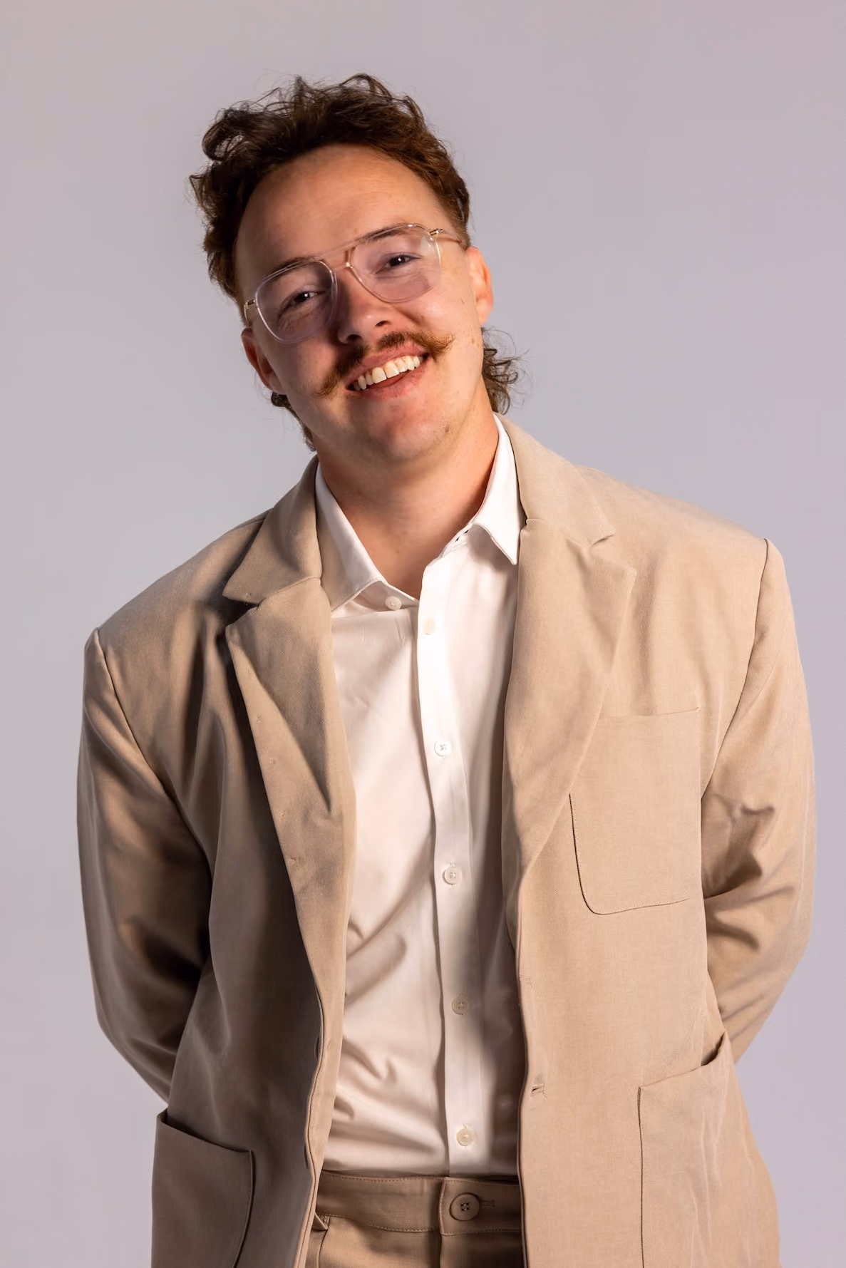 Young man with curly brown hair and mustache wearing glasses, a beige suit jacket, and a white dress shirt smiling against a plain light gray background.