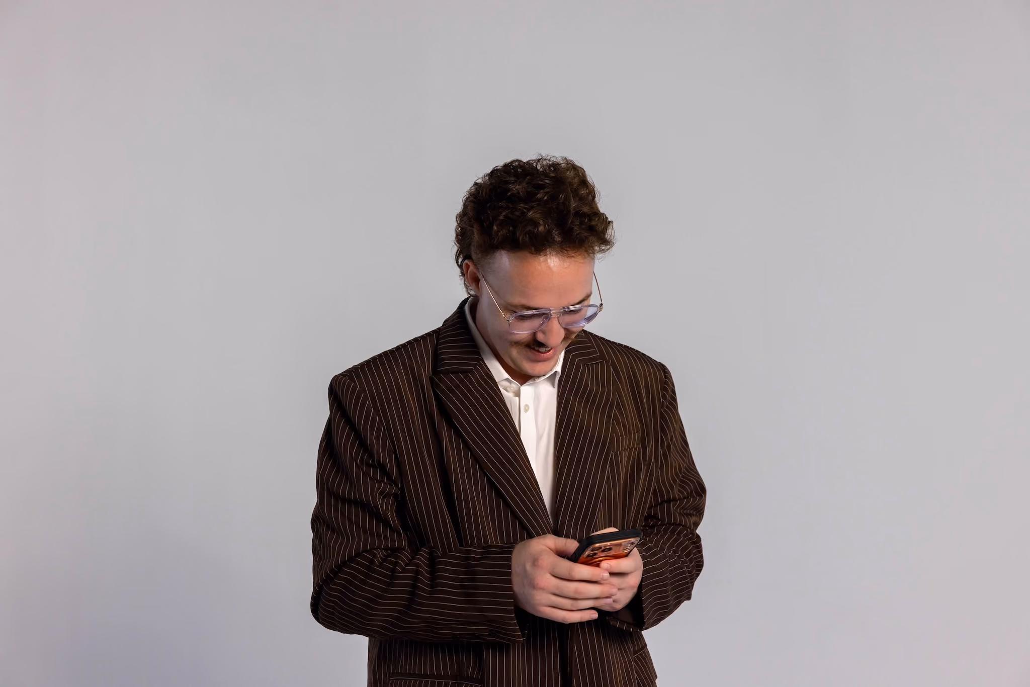 Young man with curly hair and glasses wearing a dark pinstripe blazer and white shirt looking down and smiling at his smartphone against a plain gray background.