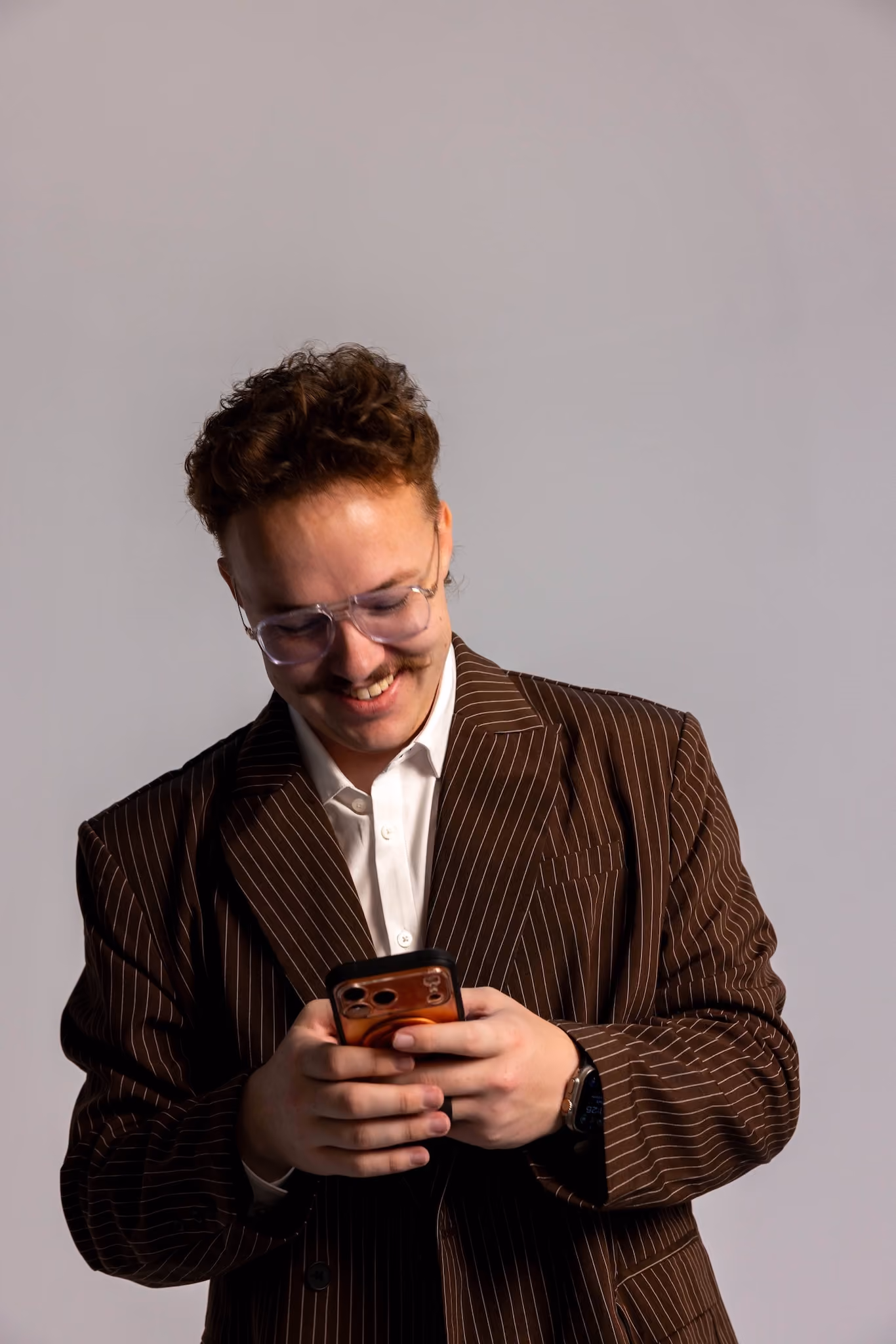Man with curly hair and mustache wearing clear glasses and a brown pinstripe suit, smiling while looking down at his smartphone.