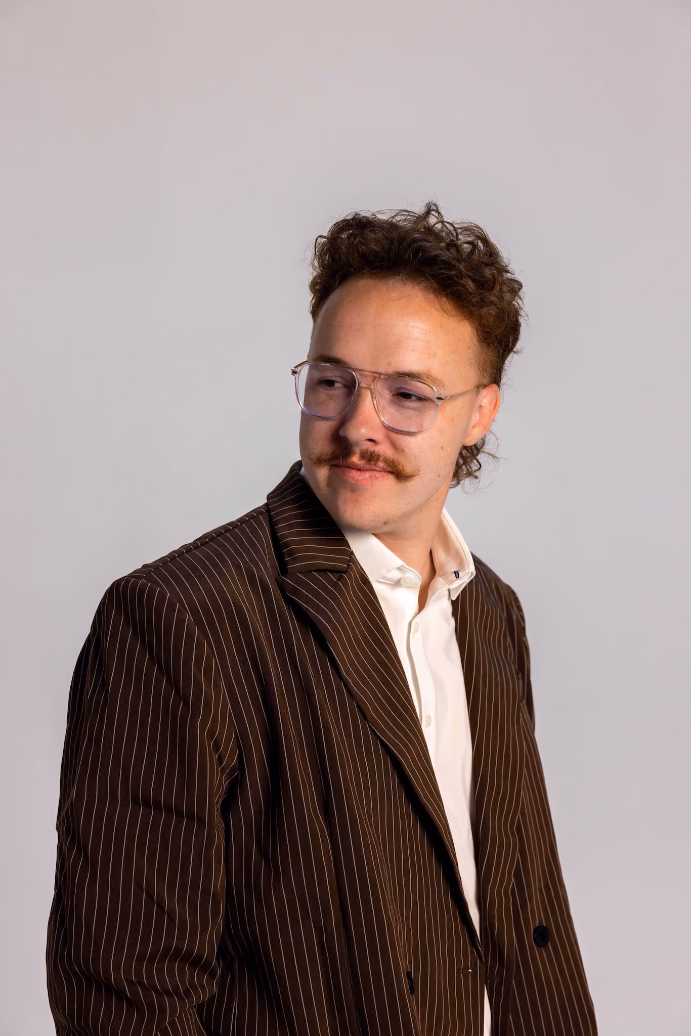 Man with curly hair and large clear-framed glasses wearing a brown pinstripe blazer and white shirt, looking to his right.