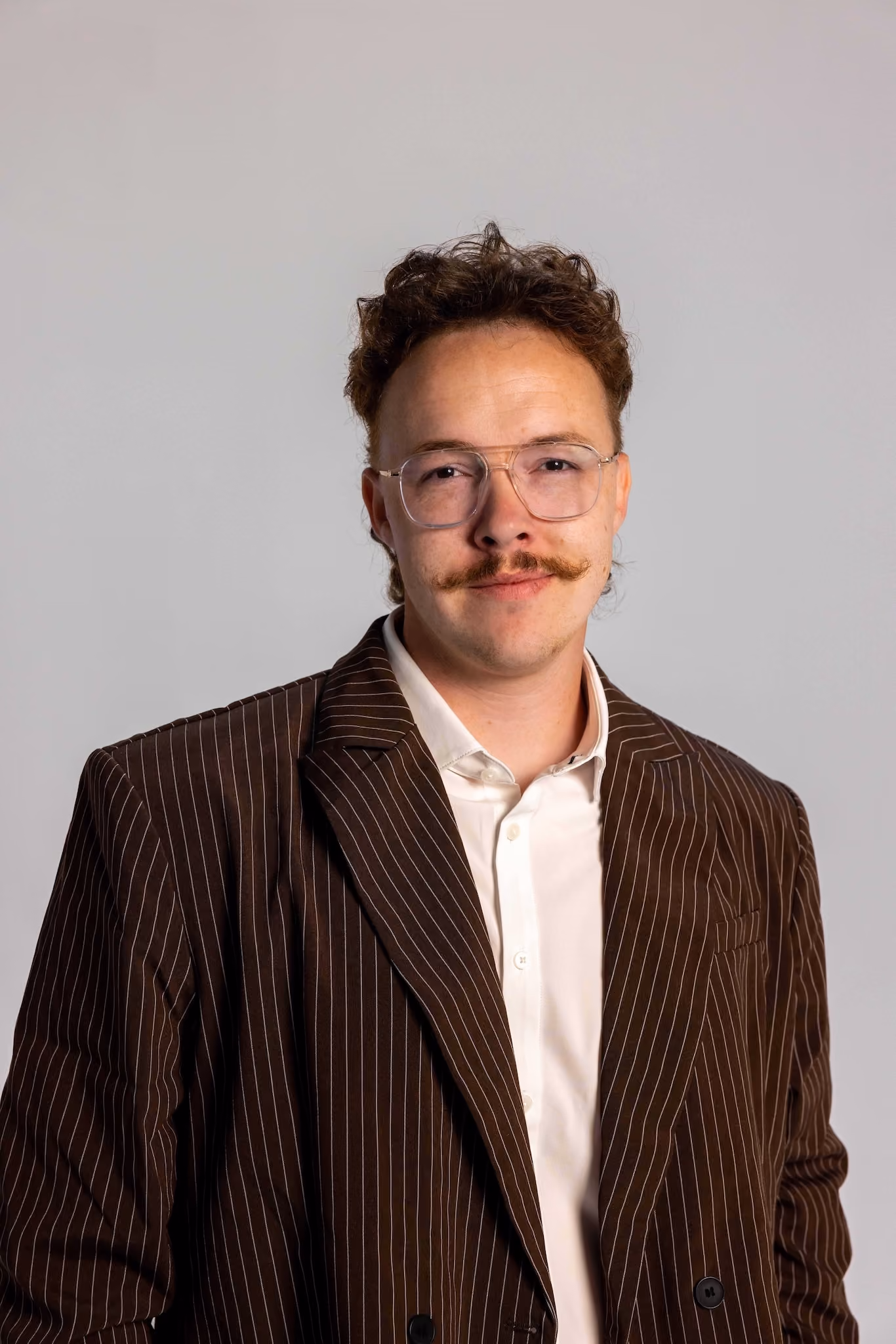 Man with curly brown hair, mustache, and clear glasses wearing a brown pinstripe blazer and white shirt, facing the camera with a neutral gray background.