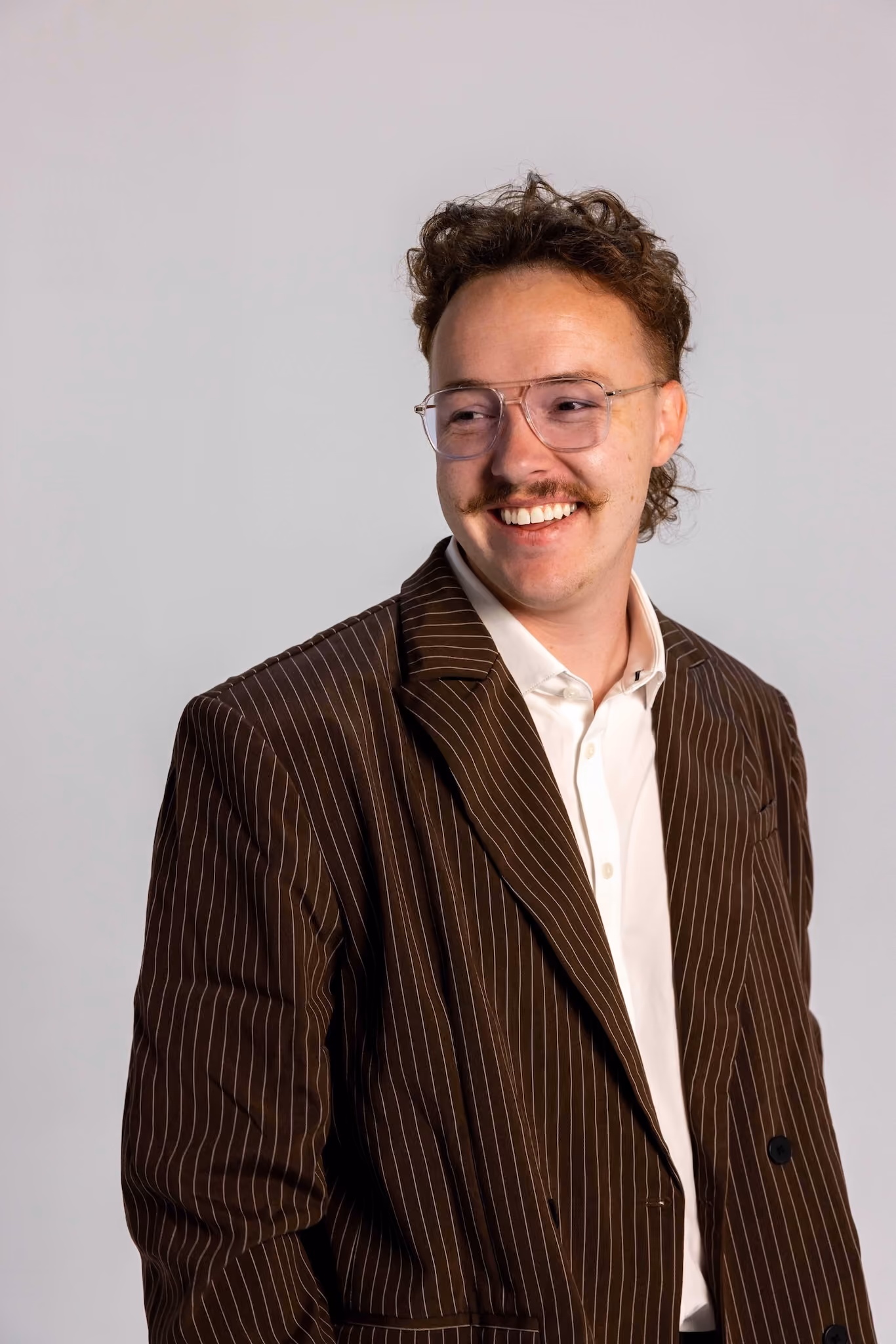 Young man with curly brown hair and mustache wearing glasses, a brown pinstripe blazer, and a white shirt, smiling and looking to the side against a plain light gray background.