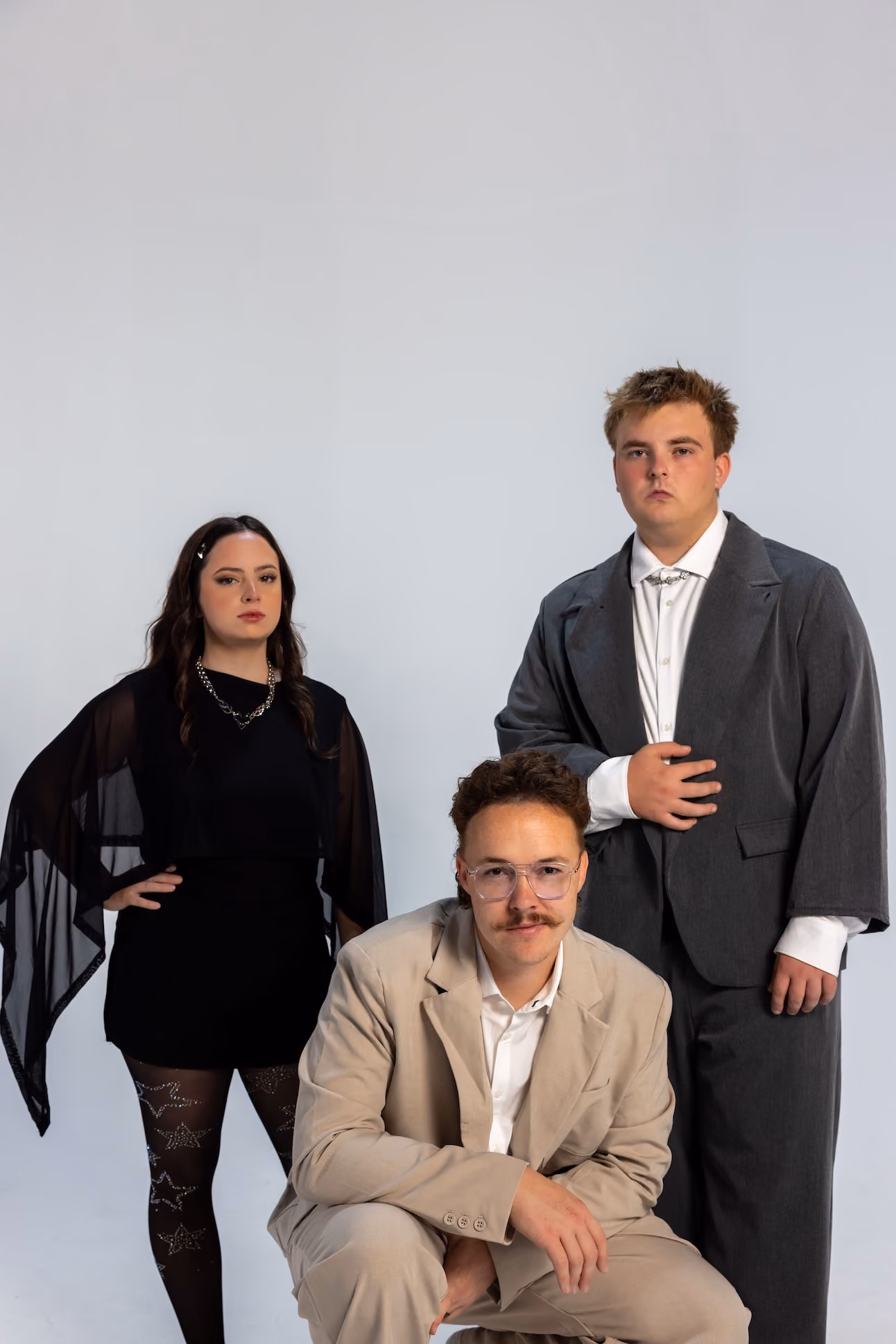 Three young adults posing against a plain background: a woman in black dress with sheer sleeves stands with hand on hip, a man in a gray oversized suit stands with one hand on chest, and another man with glasses and a mustache crouches wearing a beige suit.
