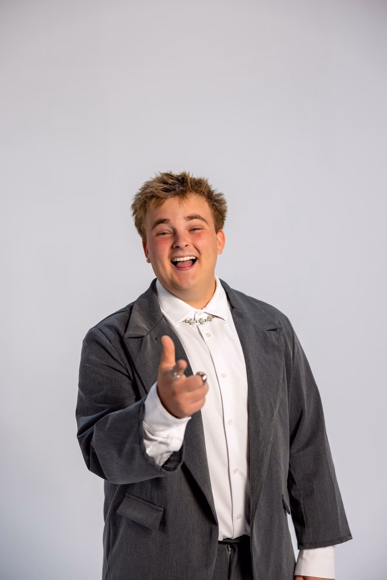 Smiling young man with short messy hair wearing a white shirt and dark gray jacket pointing forward with his right hand against a plain light gray background.