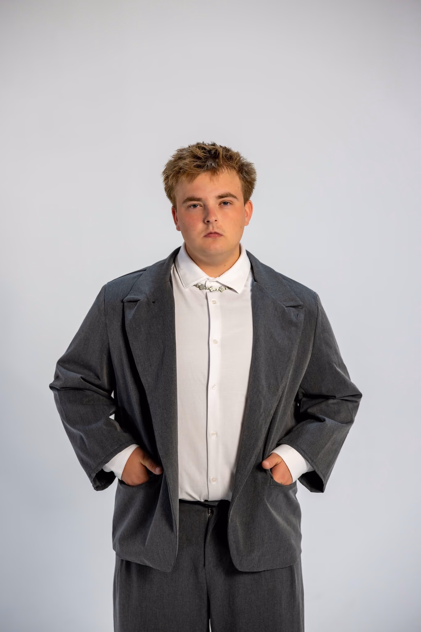 Young person with short, tousled hair wearing an oversized gray suit jacket and matching trousers over a white button-up shirt with a decorative collar chain, standing with hands in pockets against a plain light gray background.