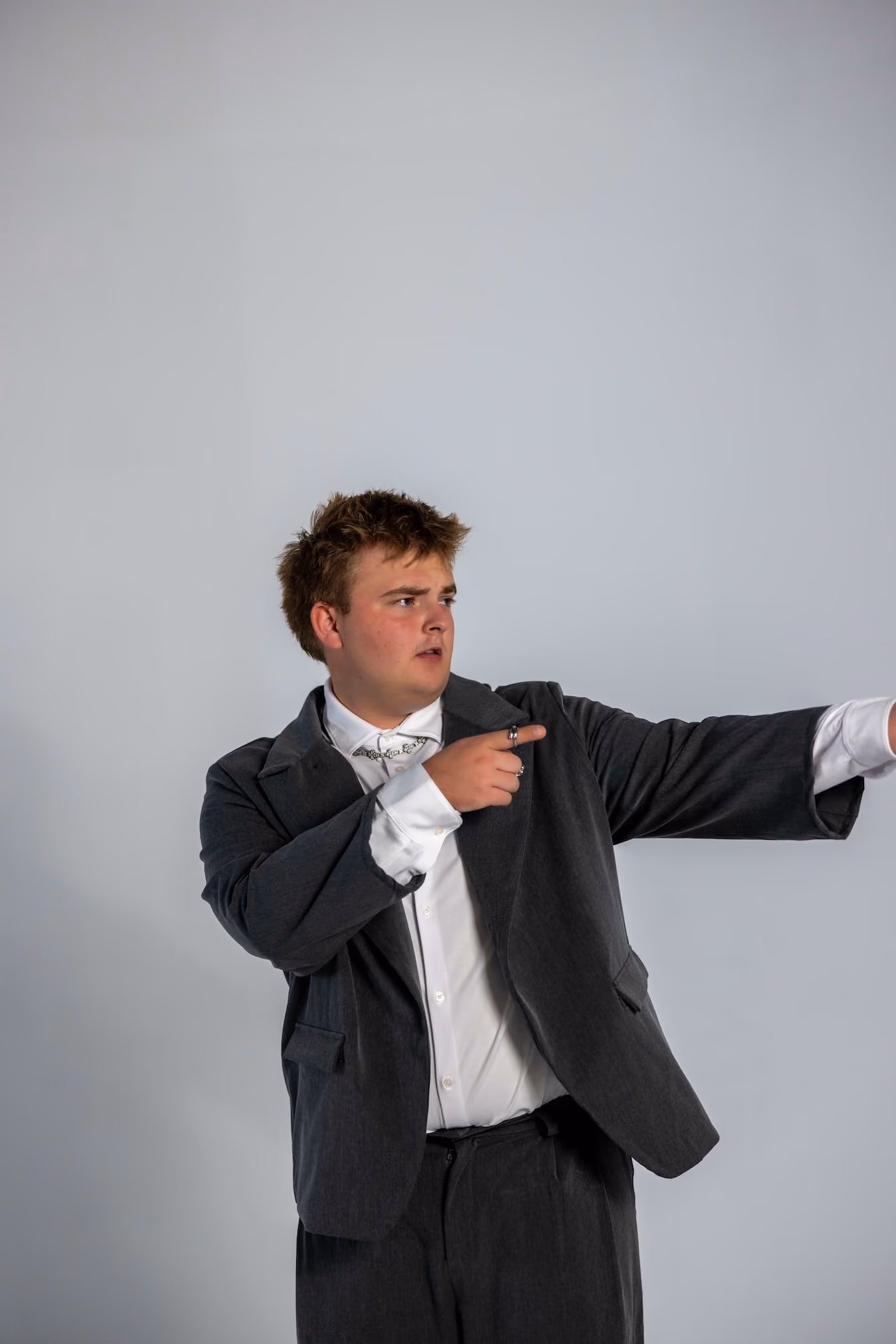 A young man with tousled hair wearing a dark suit jacket and white shirt points to his right with both hands against a plain gray background.