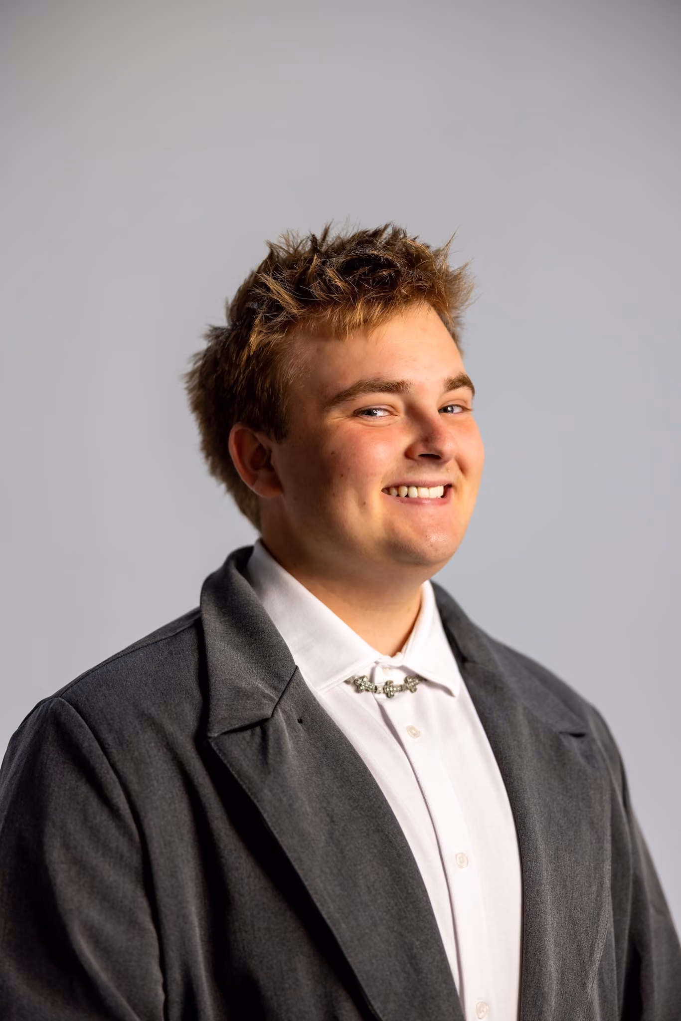 Smiling young man with short messy hair wearing a dark gray blazer over a white shirt with a decorative silver clasp at the collar, standing against a plain light gray background.
