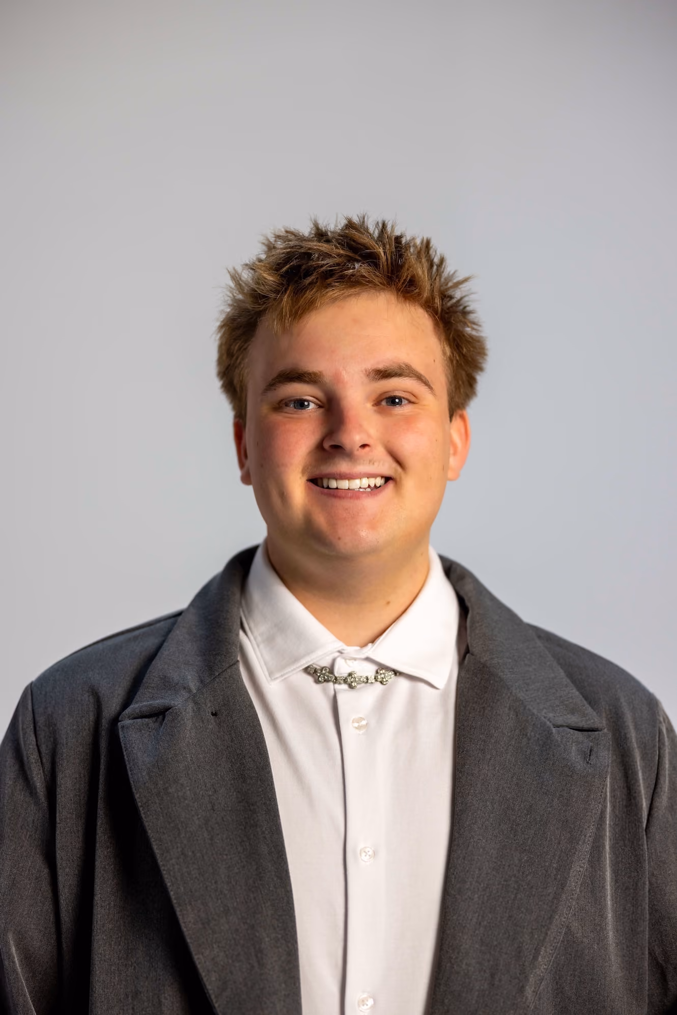 Smiling young man with light brown spiked hair wearing a white button-up shirt and a dark gray oversized blazer against a plain light gray background.