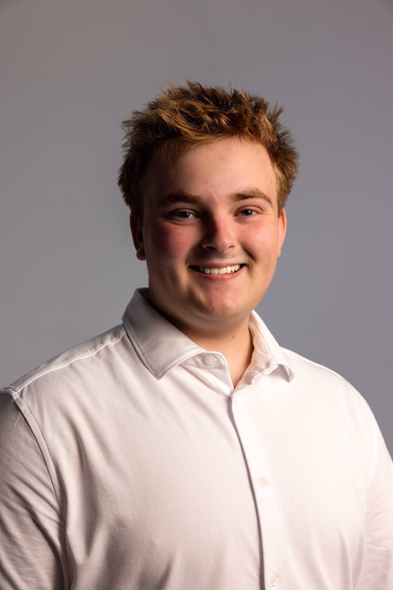 Young man with light skin and tousled brown hair smiling, wearing a white button-up shirt, against a plain gray background.