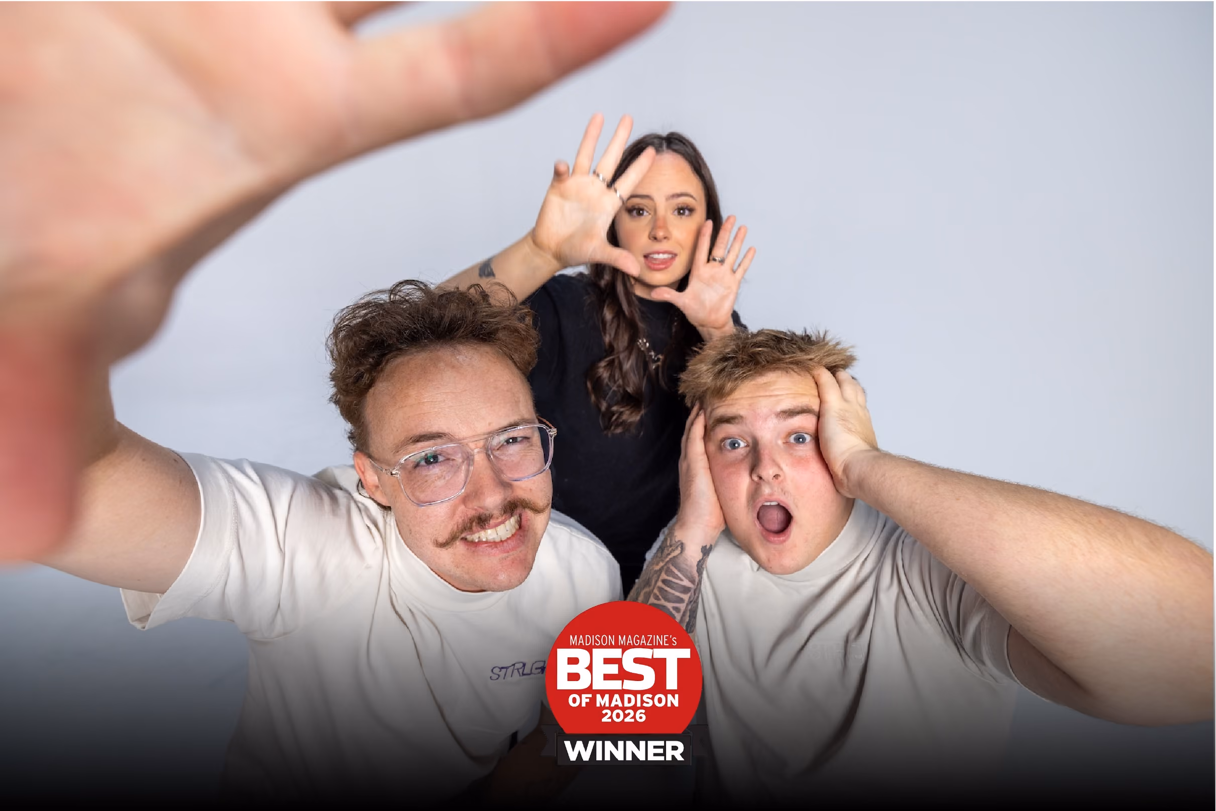 Three STRLGHT Employees posing in front of a gray background displaying a Madison Magazine's Best of Madison 2026 Winner badge; one man with glasses and mustache reaches towards the camera smiling, a woman with long hair holds her hands near her face, and another man with hands on his head looks surprised.