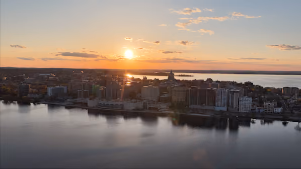 A city skyline along a waterfront at sunset with the sun low in the sky, reflecting over calm water and some scattered clouds.