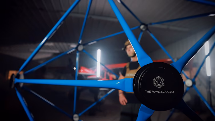 Close-up of a blue geometric exercise structure with a black center labeled 'The Maverick Gym' inside a gym, with a person wearing a workout outfit and helmet blurred in the background.