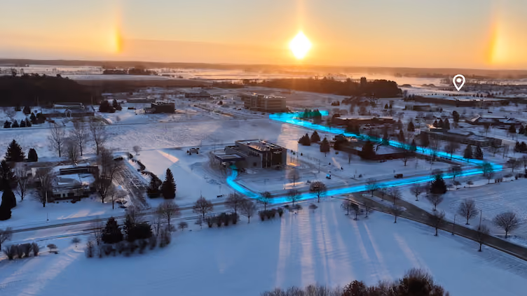 Aerial view of a snowy landscape at sunset with long shadows and a glowing sun halo causing bright light spots on either side of the sun, highlighting buildings and roads marked by a glowing blue path and a location pin icon.