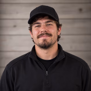Smiling man with light facial hair wearing a black cap and black quarter-zip pullover, standing against a wooden plank background.