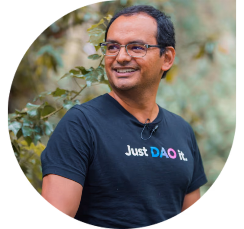 Smiling man wearing glasses and a black t-shirt that says 'Just DAO it.' standing outdoors with green leafy background.