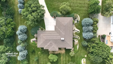 Aerial view of a house with a complex roof surrounded by a well-maintained lawn and various trees and shrubs in a landscaped yard.