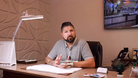 Man with a beard and mustache wearing a gray polo shirt sits at a desk with a calendar, lamp, and papers, speaking directly to the camera in an office setting.