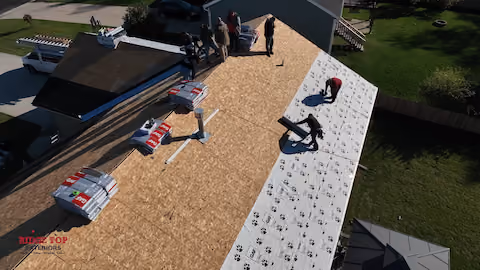 Aerial view of workers installing roofing material on a wooden roof deck, with some workers on a white underlayment and others preparing the roof surface, surrounded by green grass and neighboring roofs.