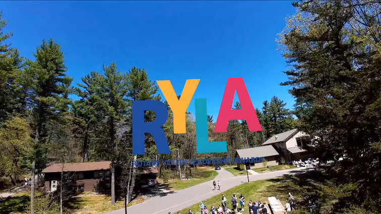 Aerial view of a wooded area with several buildings, a road, and a group of people gathering outdoors under clear blue sky, with large colorful letters RYLA overlaid on the image.