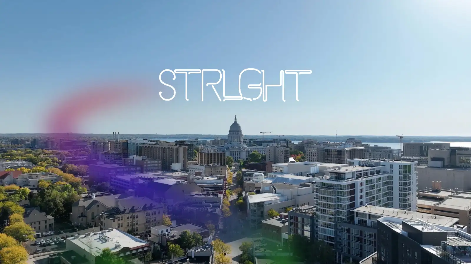A cityscape with a clear blue sky showing a prominent domed capitol building in the center and modern buildings in the foreground, with the word 'STRLGHT' in stylized white font over the sky and a faint colorful smoke effect towards the left.