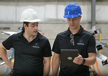 Two men wearing hard hats and black shirts looking at a tablet in an industrial setting.