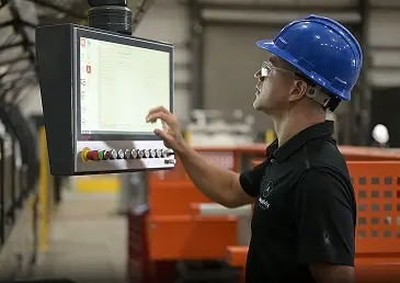 Worker wearing blue hard hat and safety glasses operating industrial touchscreen control panel.