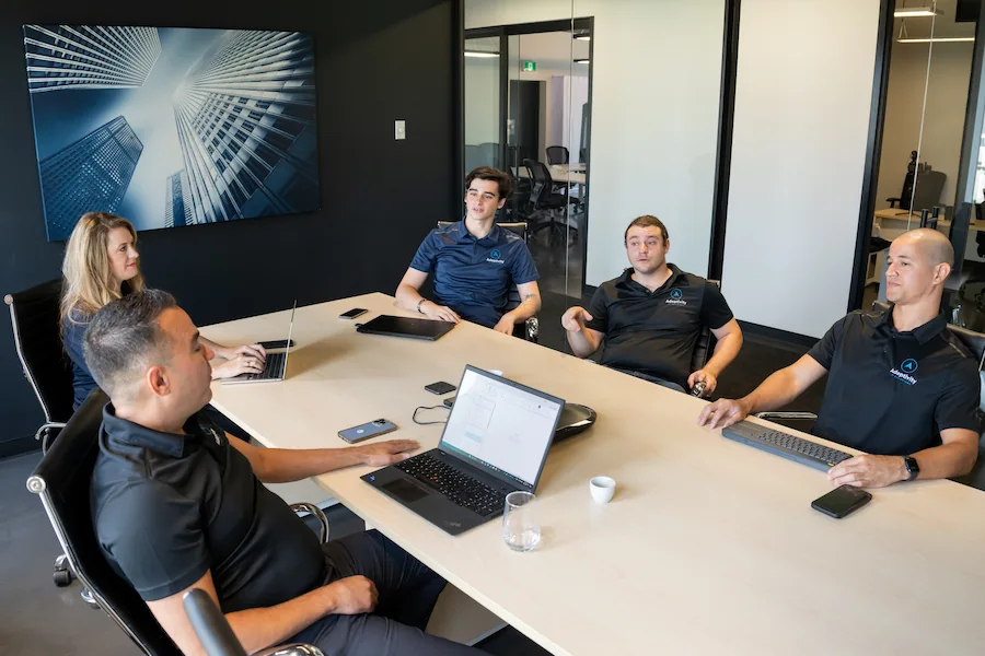 Five coworkers in matching polo shirts having a meeting around a conference table with laptops and devices.