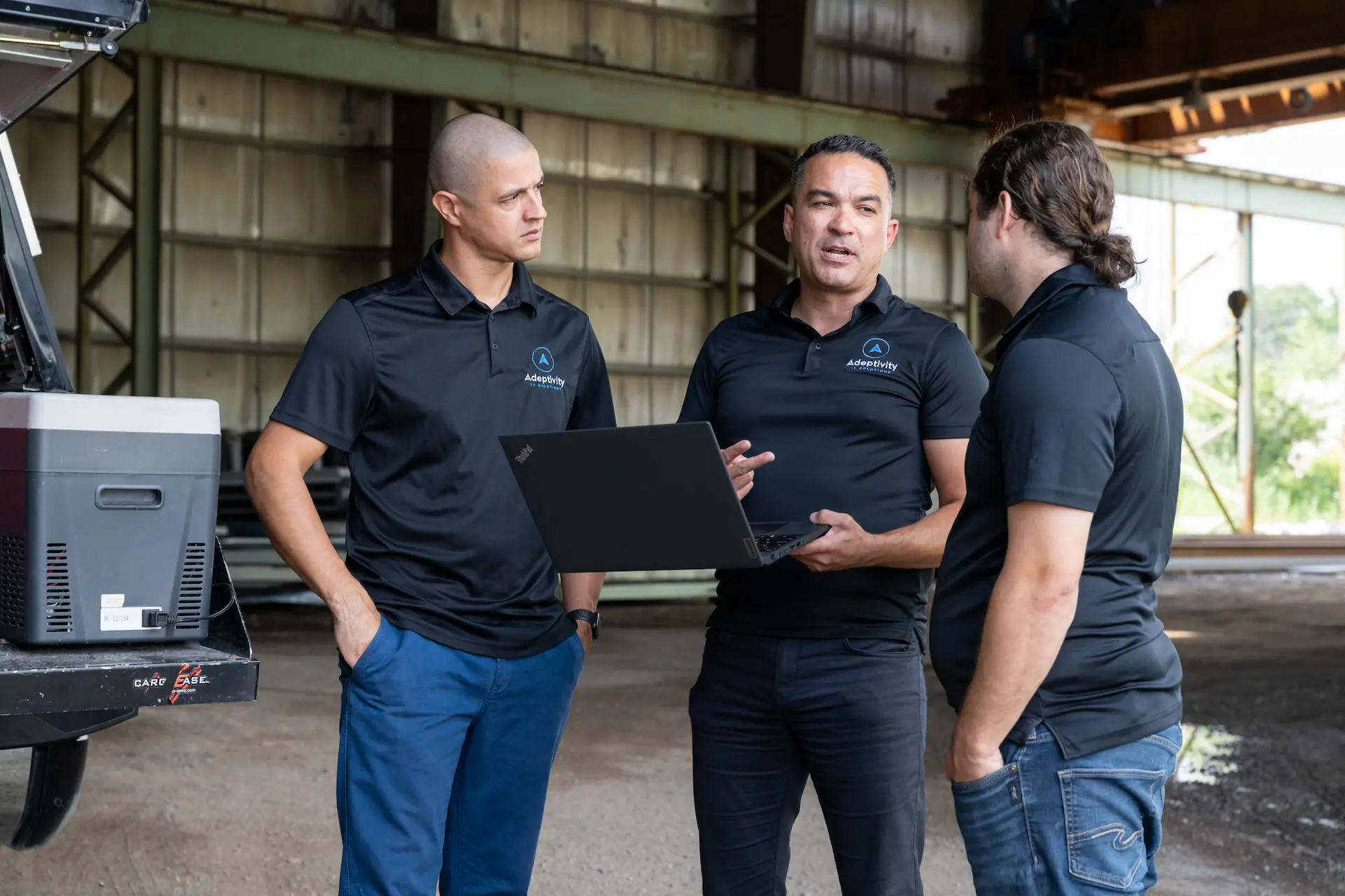 Three men wearing black polo shirts with Adeptivity IT Solutions logos stand in a warehouse-like setting, one holding a laptop while they engage in conversation.
