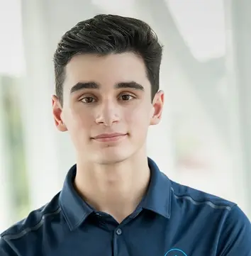 Portrait of a young man with dark hair wearing a blue collared shirt, standing indoors with a blurred background.