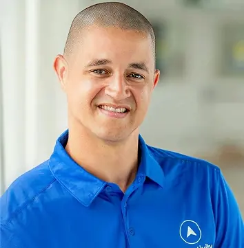 Smiling man with shaved head wearing a blue collared shirt with a white circular logo on the chest, standing indoors with a blurred background.