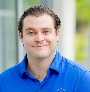 Smiling man with slicked-back dark hair wearing a blue collared shirt standing outdoors with a blurred background.