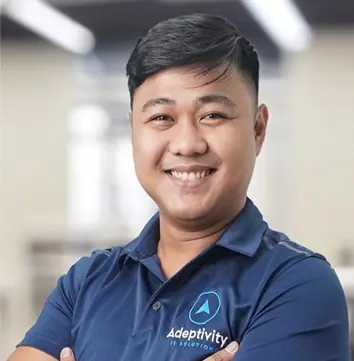 Smiling man with short black hair wearing a blue polo shirt with Adeptivity IT Solution logo, standing with arms crossed in an office setting.
