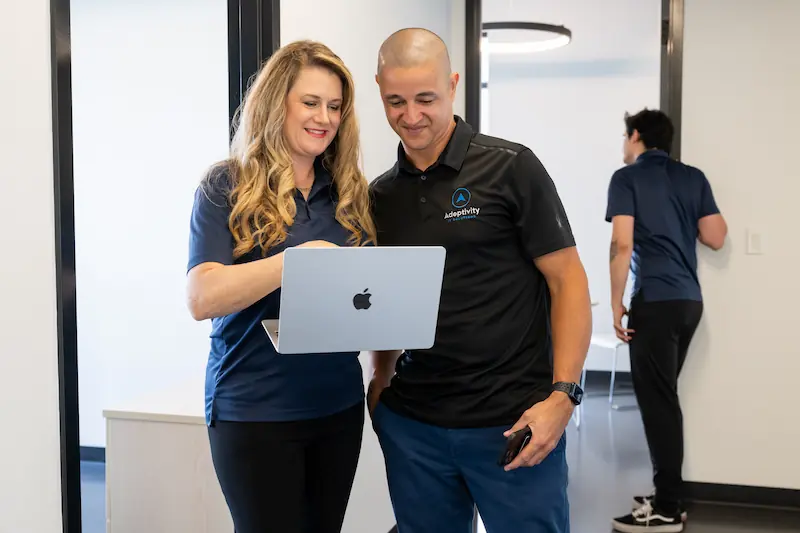 A woman and a man standing together looking at a laptop screen, both smiling, in a modern office hallway with a third person walking away in the background.