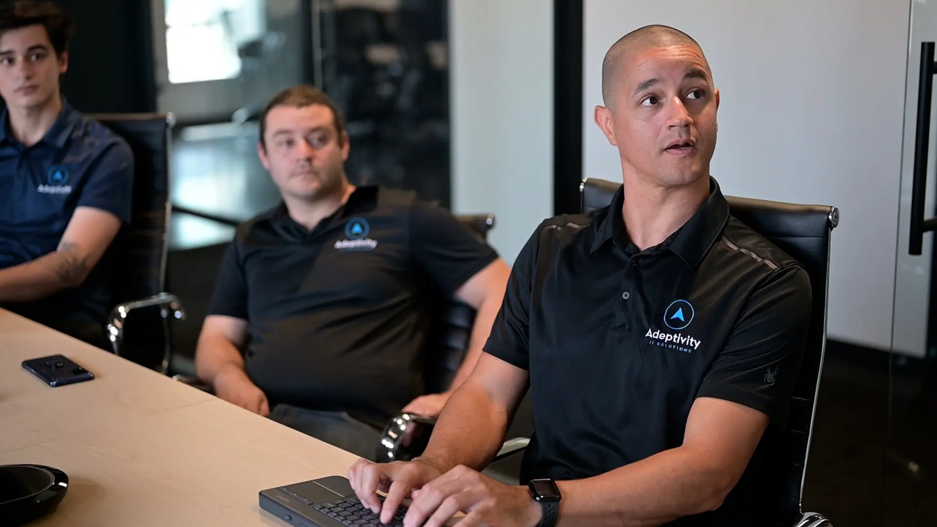 Three men sitting at a conference table in office chairs, all wearing black Adeptivity polo shirts; the man in front is typing on a keyboard and looking to the side while the other two watch attentively.