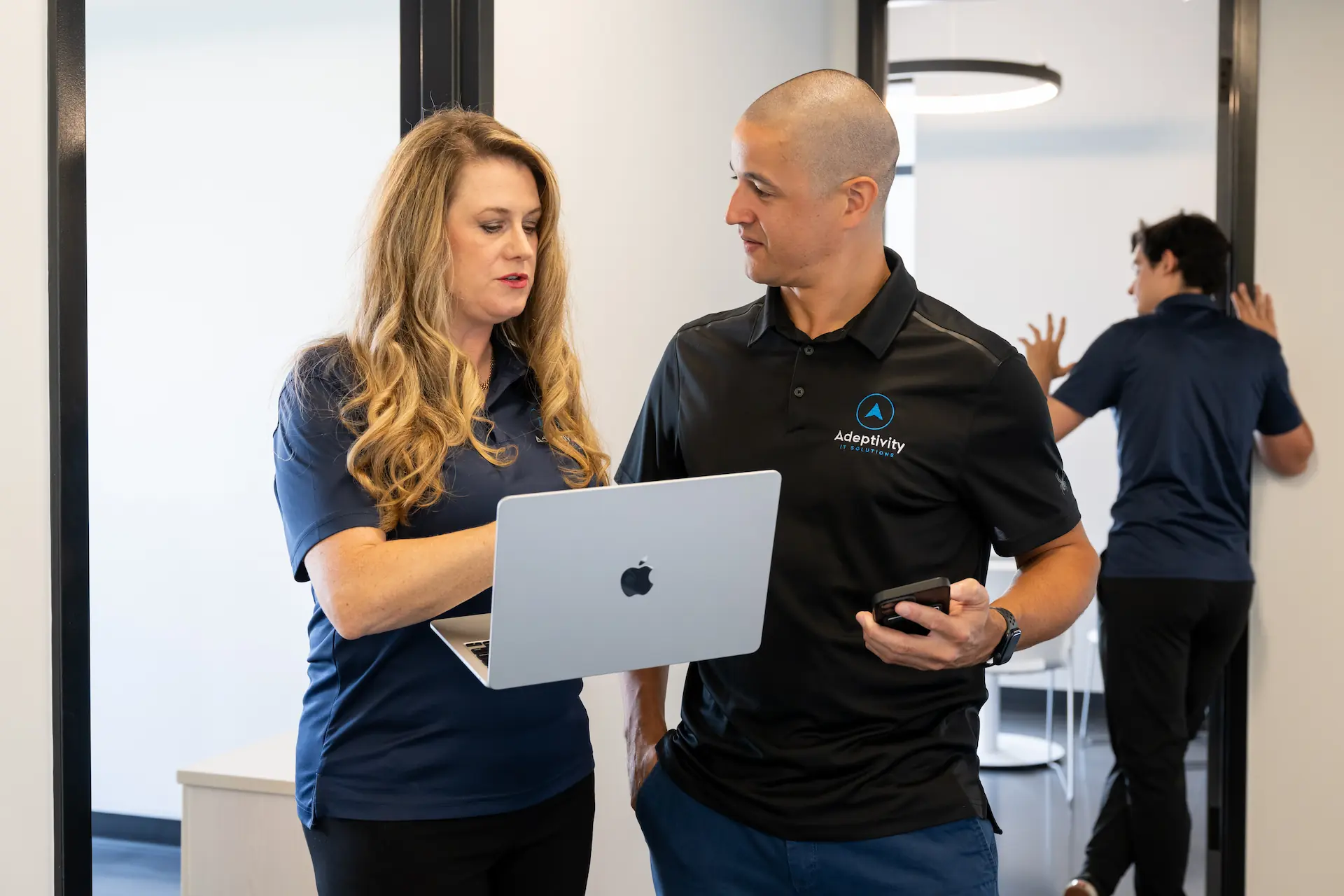 Two coworkers, a woman holding a laptop and a man holding a phone, are standing and discussing something in an office while a third person walks away in the background.