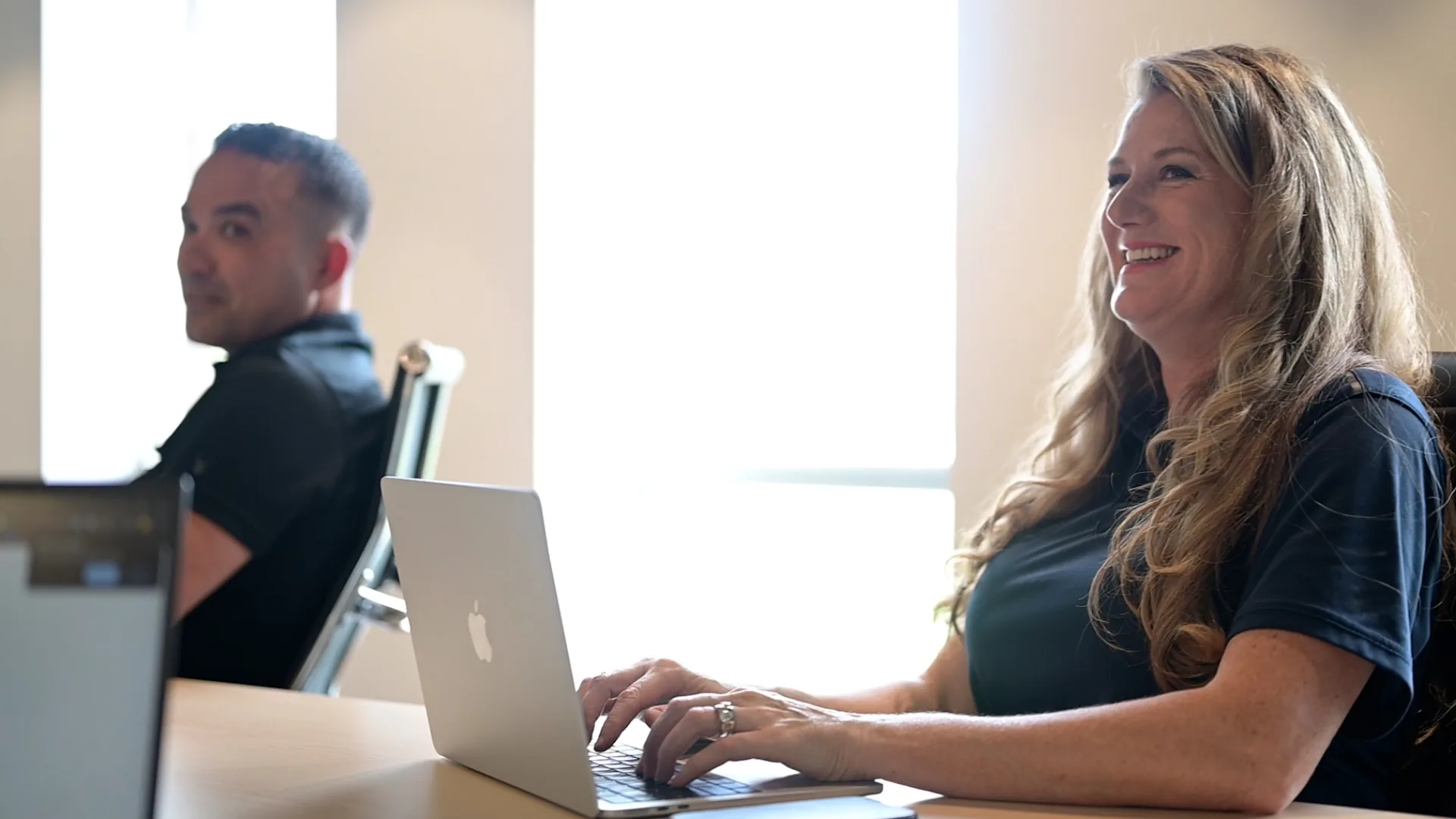 Smiling woman with long blonde hair typing on a silver MacBook laptop at a table, with a man sitting in the background.