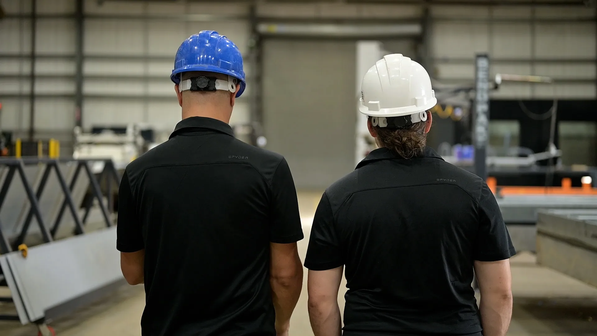 Two workers wearing black shirts and safety helmets, one blue and one white, standing with their backs facing the camera in an industrial warehouse setting.